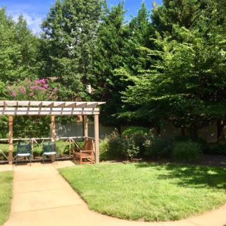 A serene garden featuring a pergola with benches, surrounded by lush green trees and a well-kept lawn.
