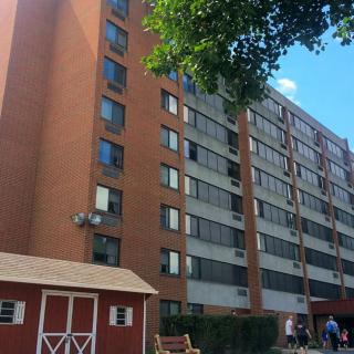 A tall apartment building with a brick facade and multiple windows, set in an urban environment with a small shed and a tree in the foreground.
