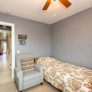 A cozy bedroom features a single bed with colorful bedding, an armchair, and a ceiling fan against gray walls.
