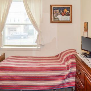 A cozy bedroom with a neatly made bed, a computer on the desk, and light filtering through curtained windows.