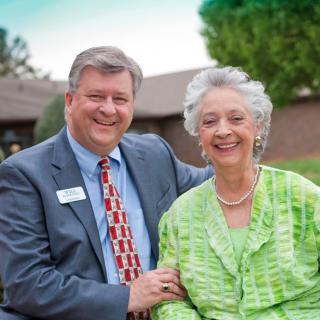 A man in a suit and an elderly woman in a green outfit are smiling outdoors.