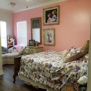 A cozy bedroom with a pink accent wall and floral bedding, featuring decorative paintings and wood flooring.
