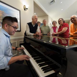 A pianist plays for an audience gathered around a piano indoors.