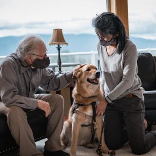 An elderly person and a woman wearing masks are petting a dog in a living room.