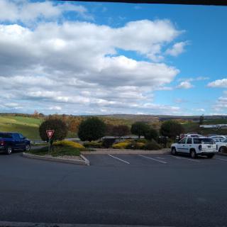 A serene landscape featuring a parking lot with vehicles, framed by lush greenery and a beautiful blue sky dotted with clouds.