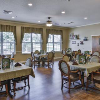 A cozy dining room with several neatly arranged tables and chairs, adorned with colorful decor and large windows allowing natural light to filter through.
