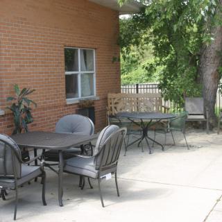 A tranquil outdoor patio area with tables and chairs beside a brick building, shaded by trees.