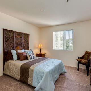 A cozy bedroom with a bed, side table, lamp, and chair next to a window.
