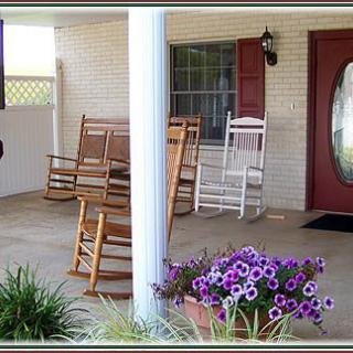 A cozy porch featuring several wooden rocking chairs, vibrant purple flowers in pots, and a welcoming front door.