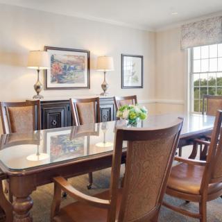 A well-lit dining room with a glass-topped table, wooden chairs, and decorative elements including artwork and a flower arrangement.