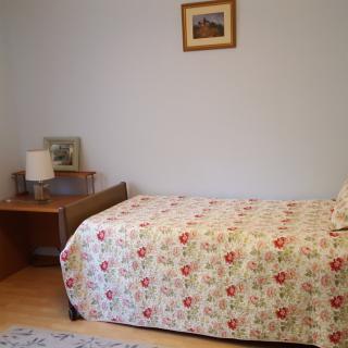 A simple bedroom with a single bed featuring a floral bedspread, beside a small desk and lamp.