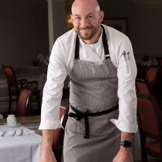 A chef standing in a restaurant setting, wearing professional attire and an apron.
