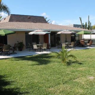 A backyard view of a house with a patio featuring tables and chairs under umbrellas, surrounded by a well-maintained lawn.