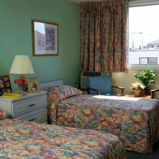 A cozy bedroom featuring two twin beds with floral patterns, a lamp on a nightstand, and curtains by the window.
