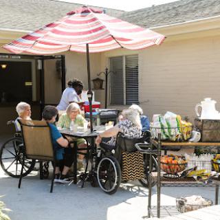A joyful gathering of seniors enjoying refreshments under a sunshade outside a care facility.
