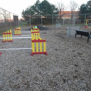 A fenced-off dog park featuring an agility course with hurdles and gravel ground.