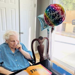 An elderly woman sitting at a table with birthday balloons and a card, celebrating her special day.