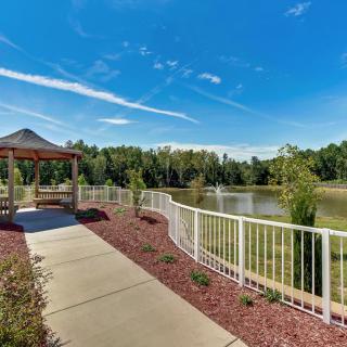 A scenic pathway leading to a gazebo by a pond, surrounded by lush trees under a clear blue sky.
