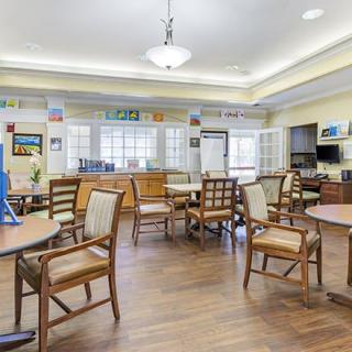 A spacious and well-lit community room with wooden floors, featuring several tables and chairs arranged for social gatherings.