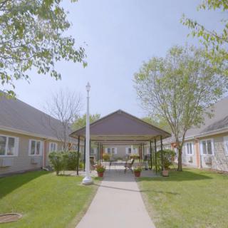 A peaceful courtyard featuring a pathway leading to a gazebo between residential buildings, surrounded by trees and greenery.