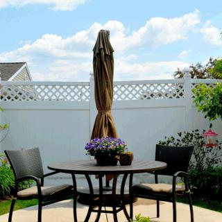 A cozy patio setup with a round table, two chairs, and an umbrella surrounded by flowers and a white fence.