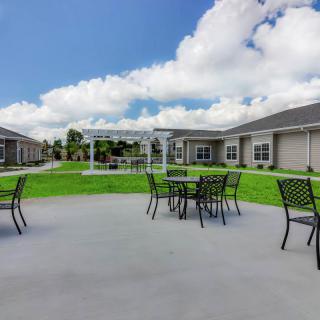 A modern patio area with tables and chairs surrounded by a green lawn and buildings under a partly cloudy sky.