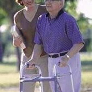 An elderly person using a walker, accompanied by a caregiver outdoors.
