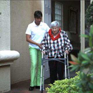 A caregiver assists an elderly person using a walker outside of a building.