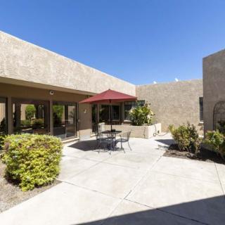 A sunny courtyard featuring a patio with a red umbrella, table, and chairs surrounded by desert plants.