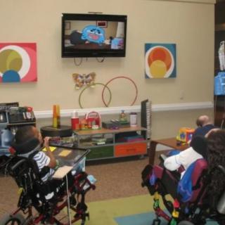 Children in wheelchairs watching television in a decorated room.