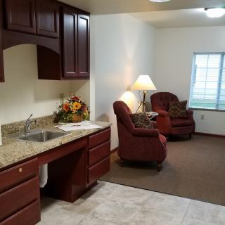 A cozy living area with a small kitchen space, featuring armchairs, a lamp, and cabinets.
