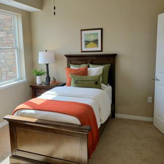 A cozy bedroom with a single bed, adorned with colorful pillows and a vibrant throw blanket. A lamp sits on the nightstand next to a window, and wall art decorates the space above the headboard.