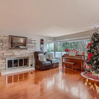 A cozy living room with a festive Christmas tree, brick fireplace, piano, and large windows.