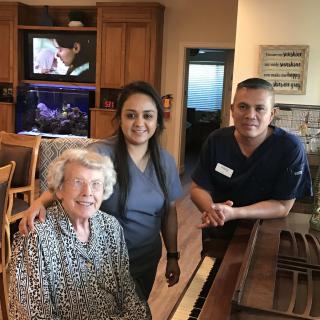 An elderly woman with two caregivers in a living room setting near a piano.