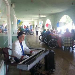 A musician playing a keyboard at an outdoor event where people are gathered, some sitting in wheelchairs and the area is decorated with balloons.