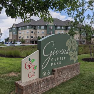 A welcoming sign for Givens Gerber Park, featuring lush greenery and a modern building in the background.