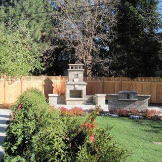 A serene garden featuring a stone fountain, patio area, and lush greenery surrounded by a wooden fence.