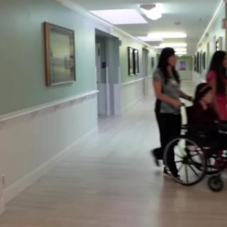 A caregiver pushes a person in a wheelchair down a hospital corridor.