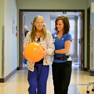A healthcare professional assists a senior patient during a rehabilitation session, using an orange ball for support.