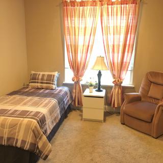 A cozy bedroom with a single bed, armchair, and a nightstand next to a window with bright curtains.
