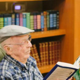 An elderly man wearing a cap is reading a book in a library setting, surrounded by shelves of books.
