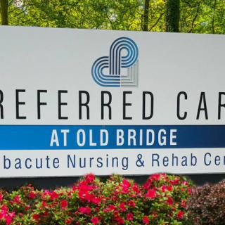A sign for Preferred Care at Old Bridge, a subacute nursing and rehab center, surrounded by flowers.