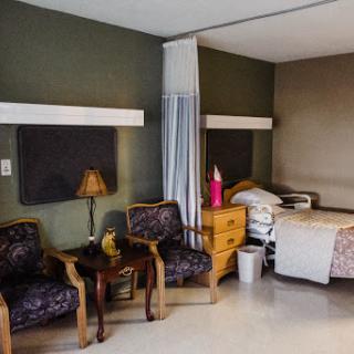 A cozy patient room featuring a bed, two chairs, a lamp, and decorative elements.
