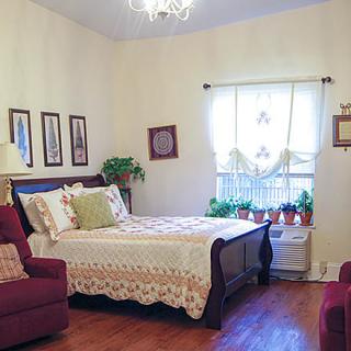 A cozy bedroom with classic decor, featuring a bed with patterned bedding, artwork on the walls, and potted plants by the window.