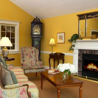 A cozy living room with a warm yellow wall, featuring a fireplace, antique clock, and comfortable seating.