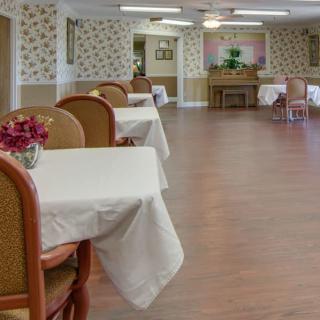A well-arranged dining area with tables covered in white tablecloths and flowers on each table, set in a room with wallpaper and wood flooring.