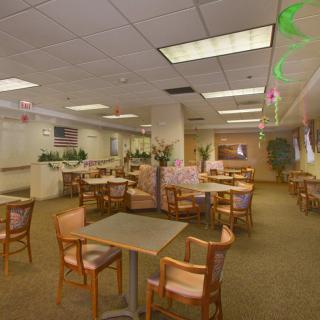An empty cafeteria with tables and chairs arranged neatly, surrounded by potted plants and illuminated by ceiling lights.