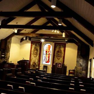 Interior view of a church featuring wooden beams, pews, and a stained glass window.