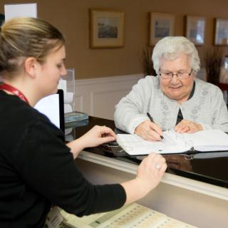 An elderly woman signing documents at a reception desk.