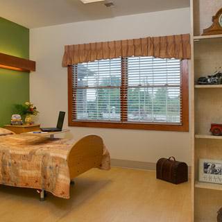 A cozy hospital room featuring a single bed, large window with blinds, and shelves for personal items.